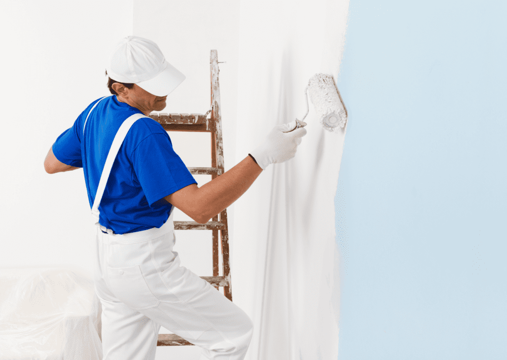 A painter in a white and blue uniform uses a roller to paint a wall from light blue to white, standing next to a ladder.