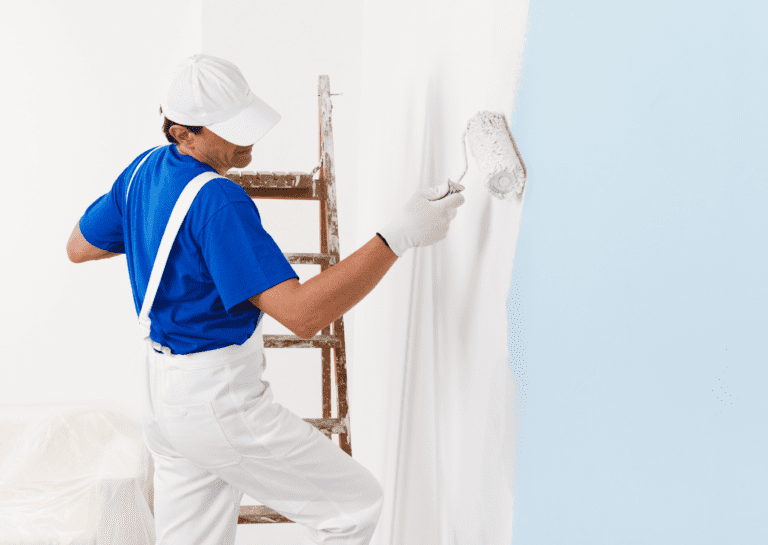 A painter in white and a blue uniform uses a roller to paint a wall from light blue to white, standing next to a ladder.