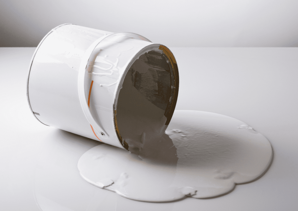 A white paint can be tipped over, spilling its contents onto a smooth white surface, creating a large puddle of paint.