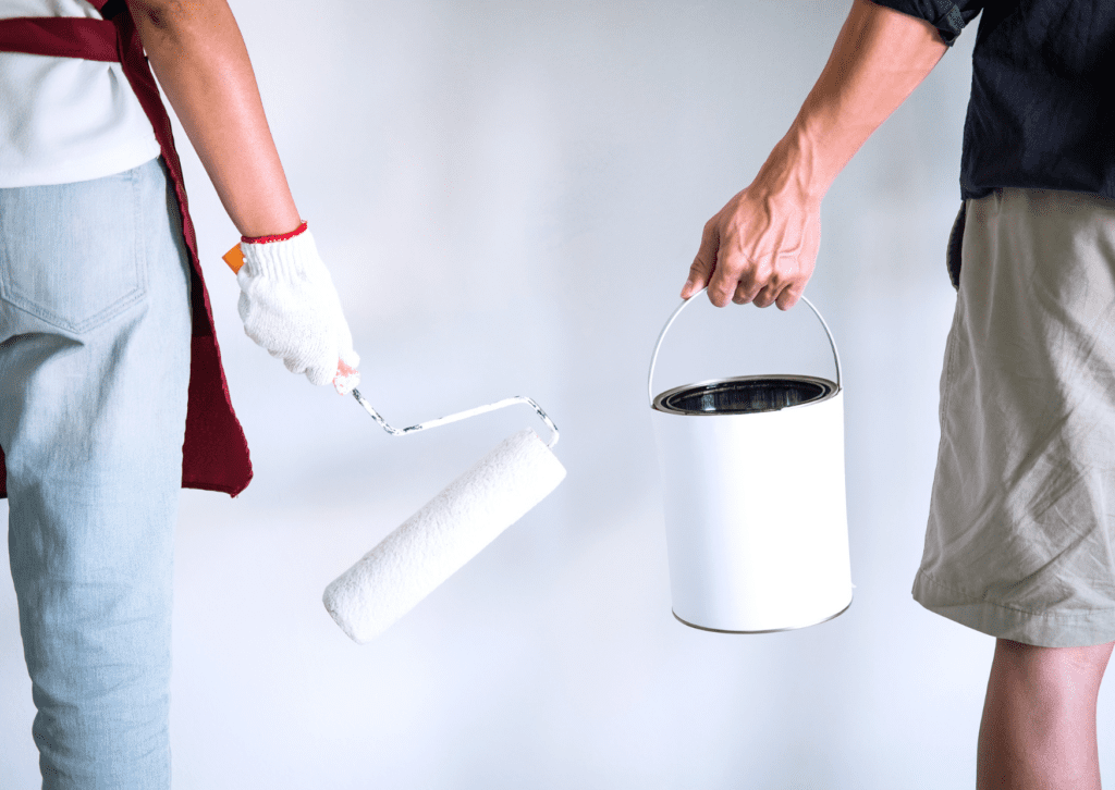 Two people stand side by side, one holding a paint roller and the other holding a white paint can, ready for painting.