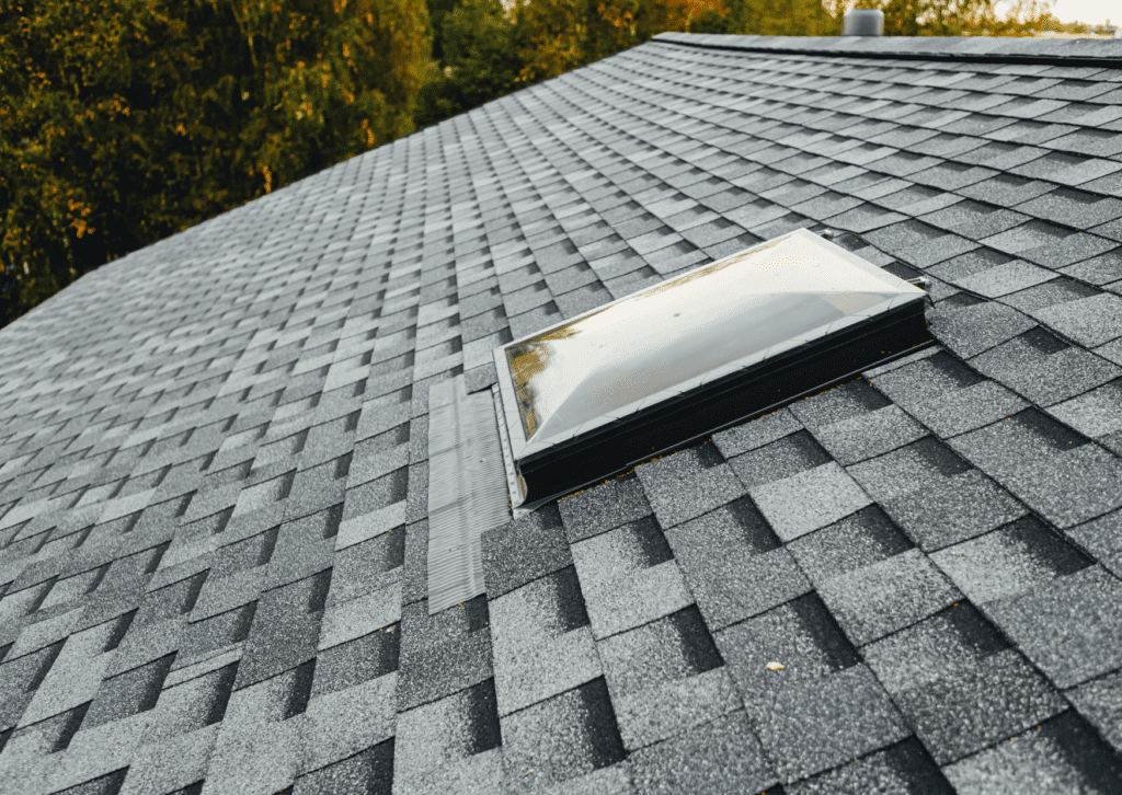 A sloped roof with gray shingles and a skylight, showcasing high-quality roofing materials surrounded by green trees.