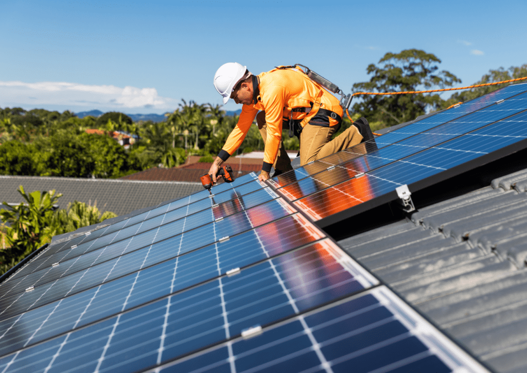 Worker in safety gear and a white hard hat installing solar panels on a rooftop with a drill, surrounded by lush greenery.