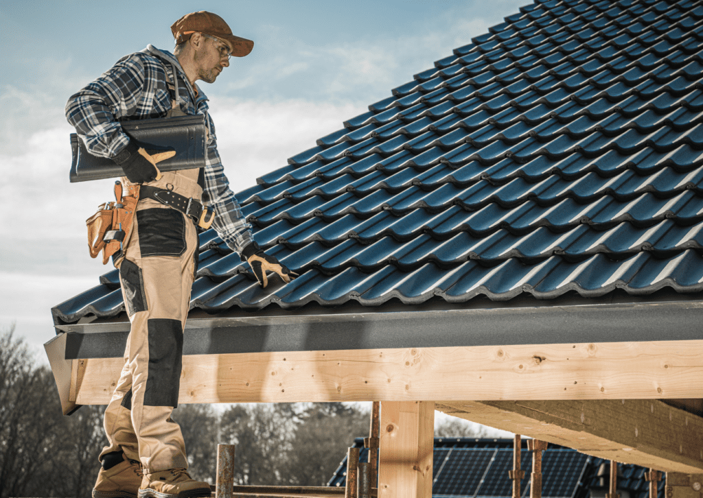 A roofer in a plaid shirt and tool belt inspecting newly installed black roof tiles on a wooden house under a clear sky.