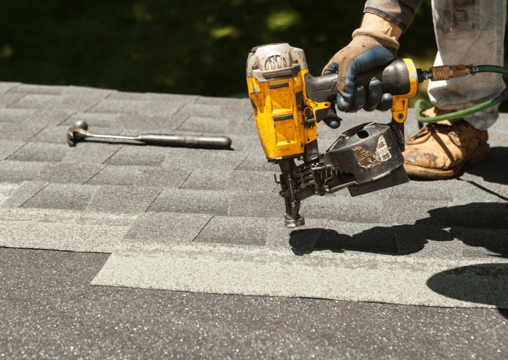 Close-up of a worker using a nail gun to install asphalt shingles on a roof, with a hammer and tools visible on the shingles.