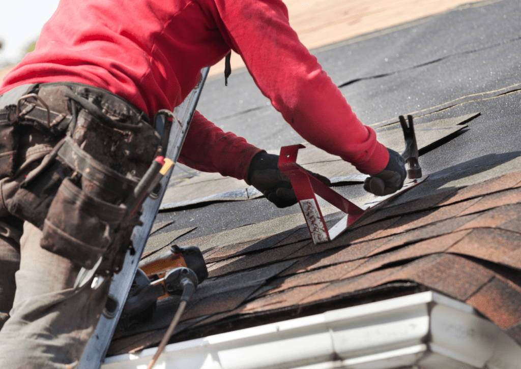 A roofer in a red shirt used tools to install shingles on a roof, with a tool belt around his waist and a ladder nearby.