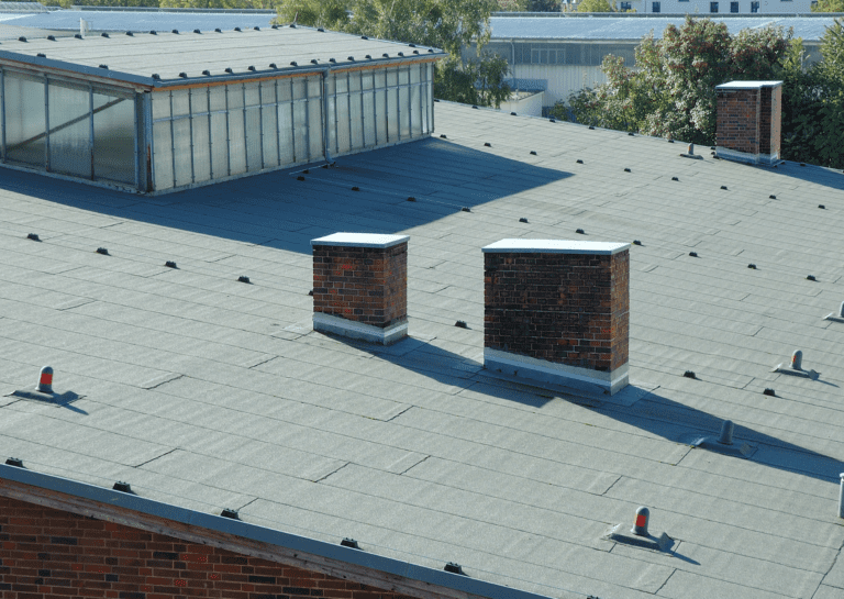 A flat rooftop with gray shingles, featuring brick chimneys and skylights, surrounded by greenery and industrial buildings.