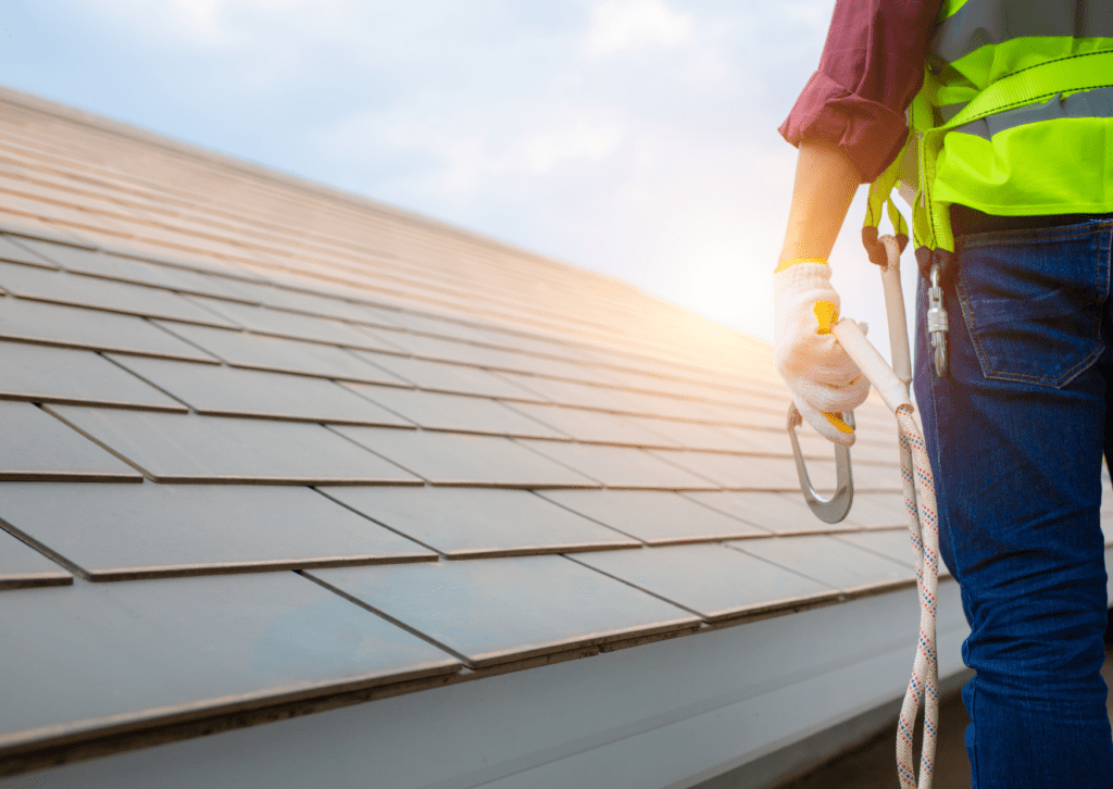 A worker in a safety vest and gloves stands on a rooftop with a safety harness, inspecting the shingles as the sun sets.