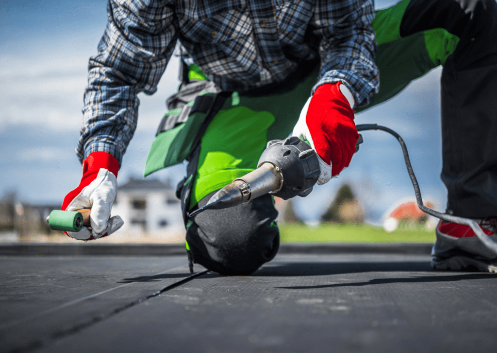 Worker in red gloves and green pants using a heat gun and roller to apply roofing membrane on a flat roof, close-up shot.