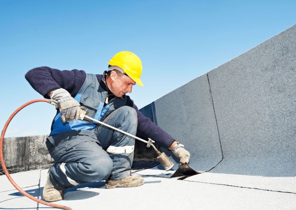 A worker in a yellow hard hat uses a blowtorch to seal a flat roof with a waterproofing membrane under a clear blue sky.