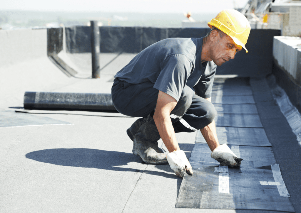 A worker in a yellow hard hat and gloves installing a waterproofing membrane on a flat roof, focusing intently on his task.