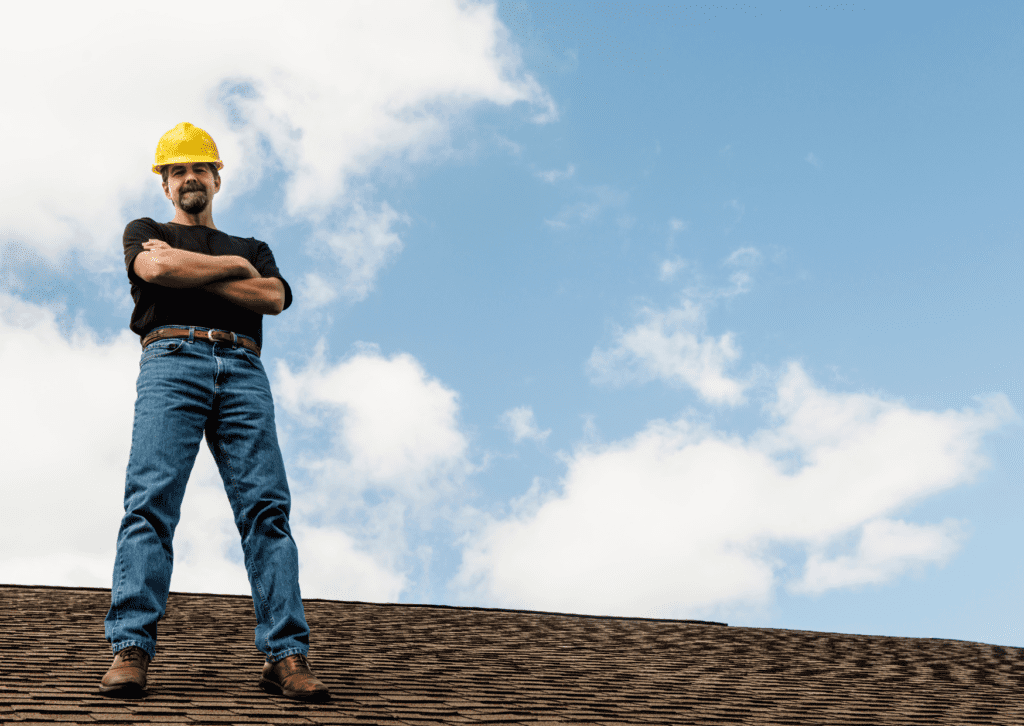 A worker in a yellow hard hat stands on a roof with arms crossed, representing top-tier roofing solutions against a blue sky