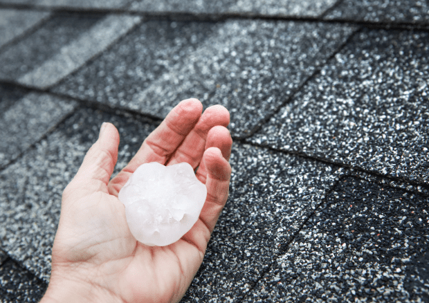 Close-up of a hand holding a large hailstone over a hail-damaged roof, highlighting the need for roof repairs due to weather.
