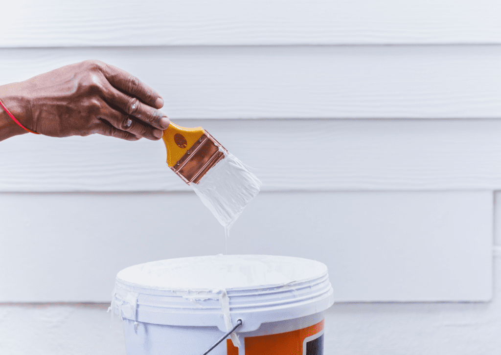 A painter dips a brush into white paint, preparing to coat the exterior siding of a house on a bright, clear day.