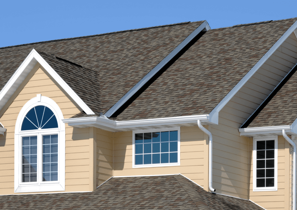 A beige house with a complex roofline featuring brown shingles and white gutters under a clear blue sky.