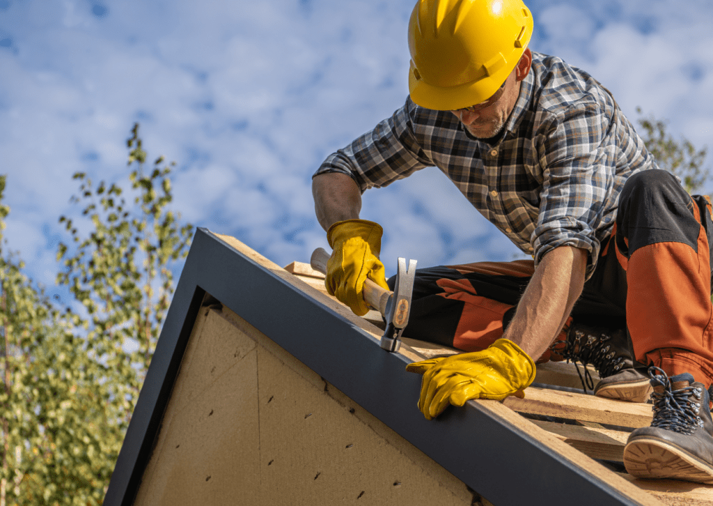 Worker providing roofing services, hammering nails into a roof, wearing protective gear on a sunny day.