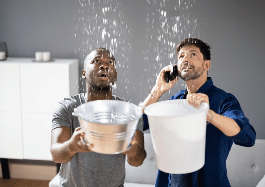 Two men catching water from roof leaks in buckets while calling for emergency repair services to address a leaking ceiling.