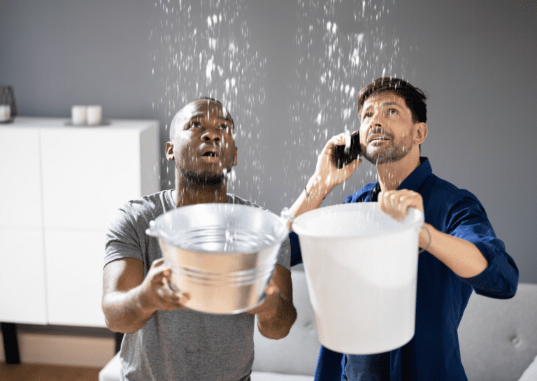 Two men catching water from roof leaks in buckets while calling for emergency repair services to address a leaking ceiling.