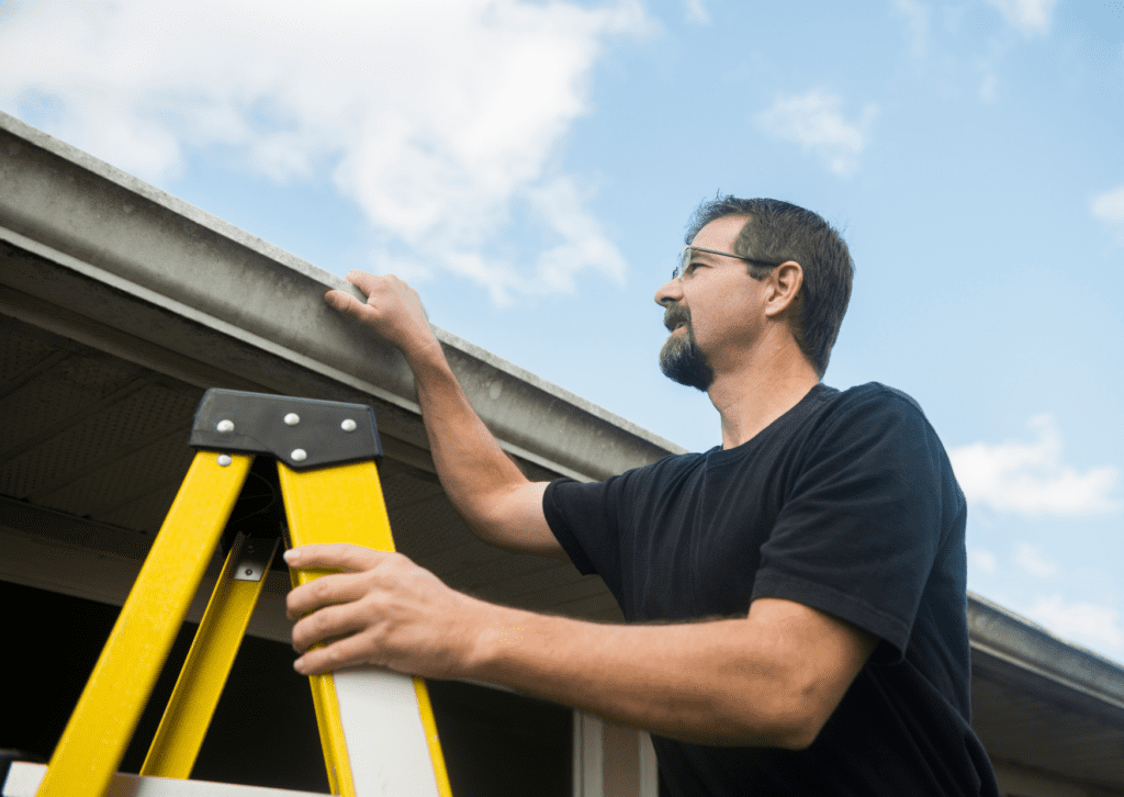 Man inspecting roof gutters from a ladder, performing routine roof maintenance and gutter cleaning for home upkeep.