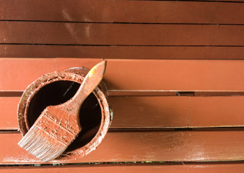 Top view of a paintbrush resting on an open can of brown wood paint, with freshly painted wooden slats in the background.