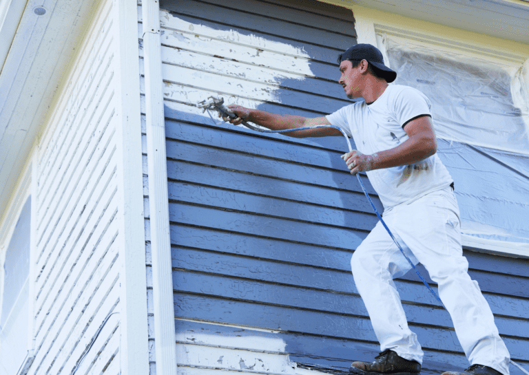 Professional painter using a sprayer to paint house siding, demonstrating ideal weather for painting outside.