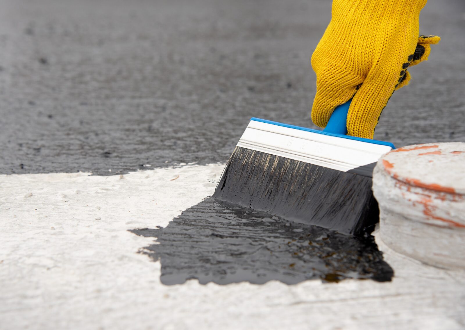 A worker in yellow gloves is applying black sealant to a flat surface with a wide brush for waterproofing.