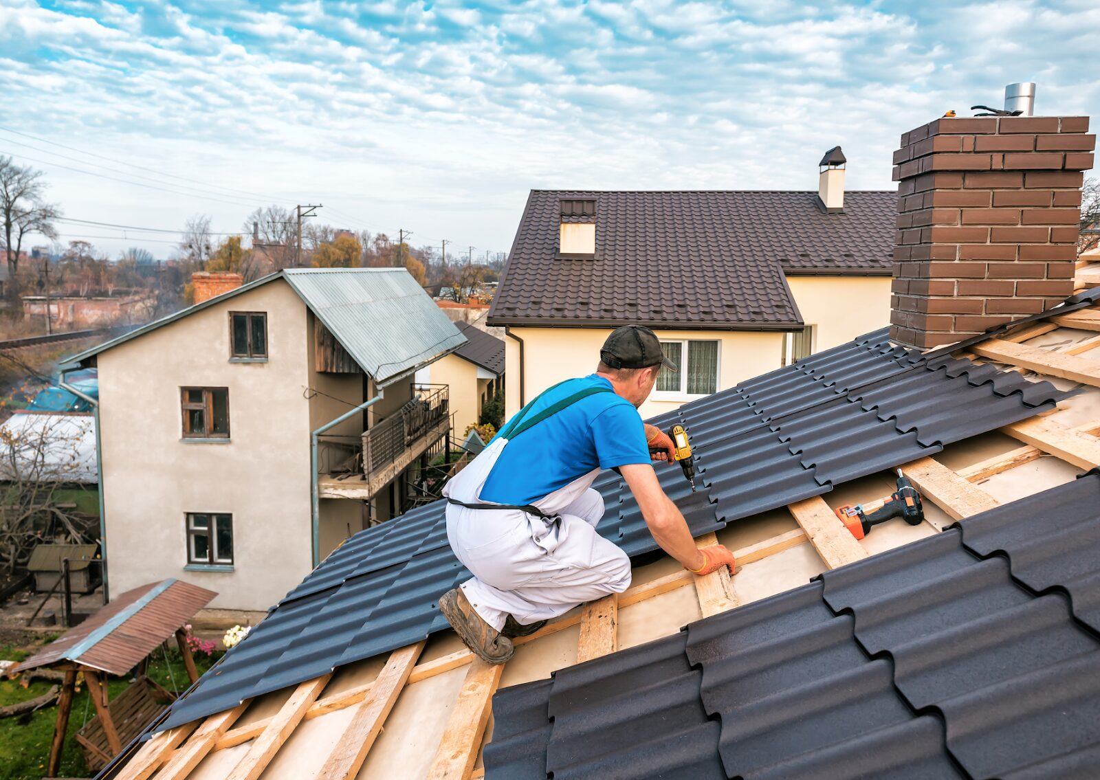 Roofer installing black metal roofing sheets on a residential house roof using a drill under a blue sky.