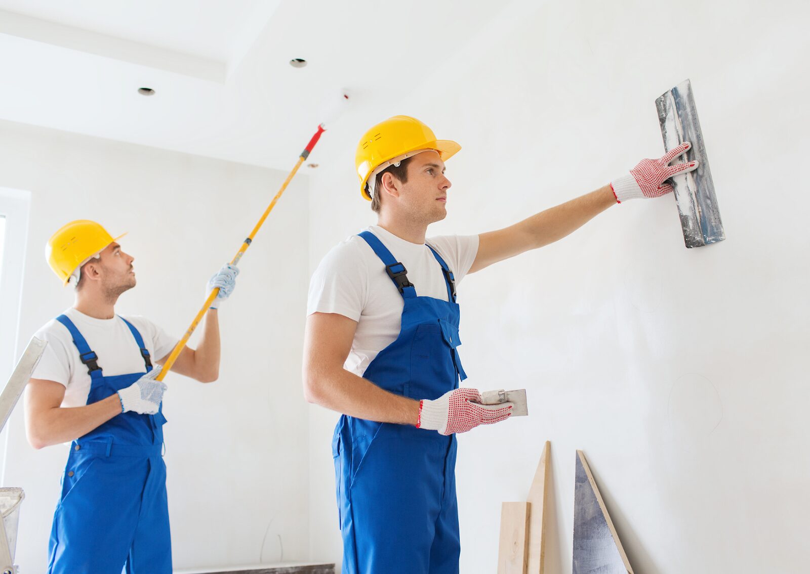 Two workers in blue overalls and yellow helmets are painting and smoothing a white interior wall during renovation.