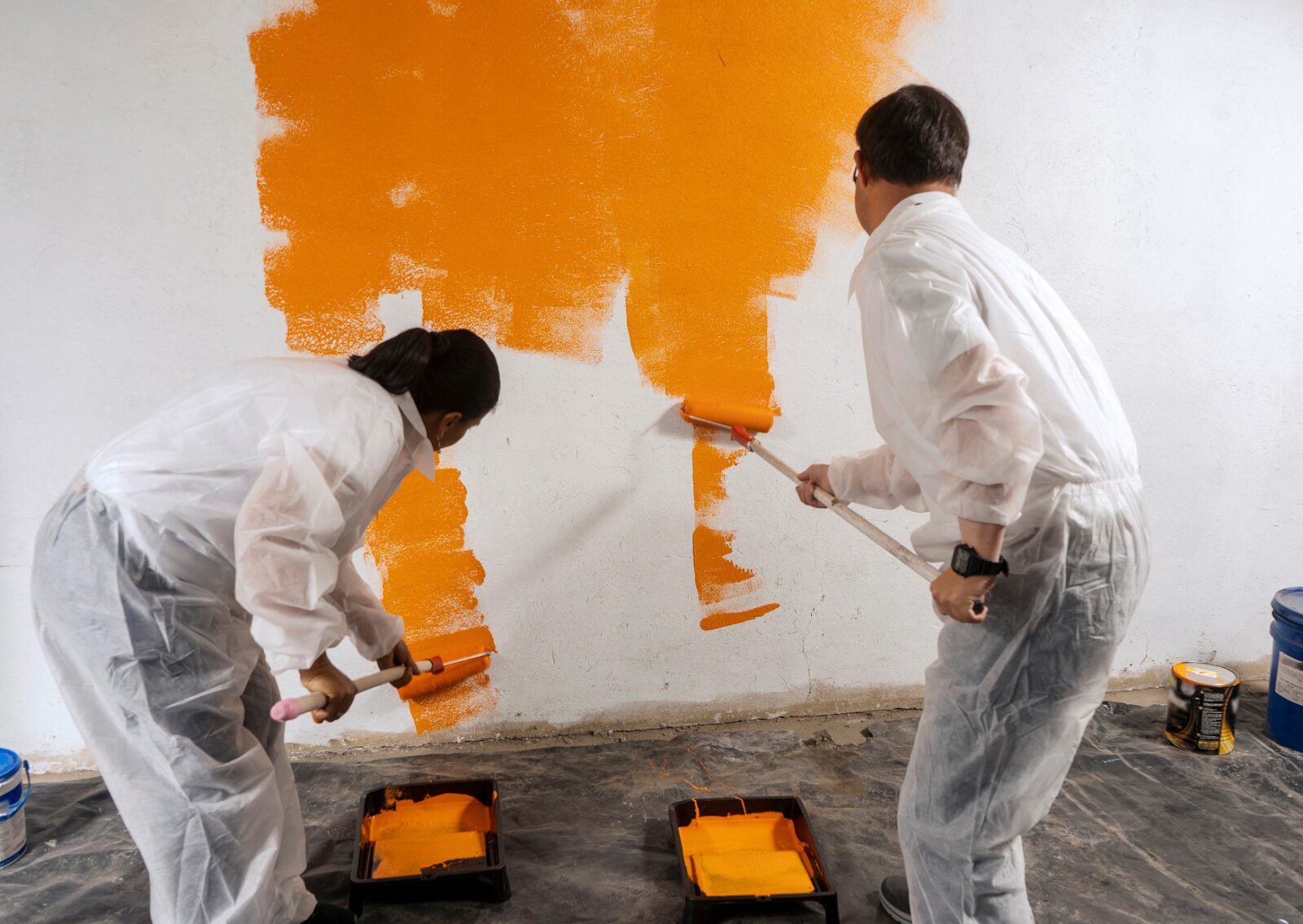 Two painters in protective suits are using rollers to apply bright orange paint to a white interior wall.