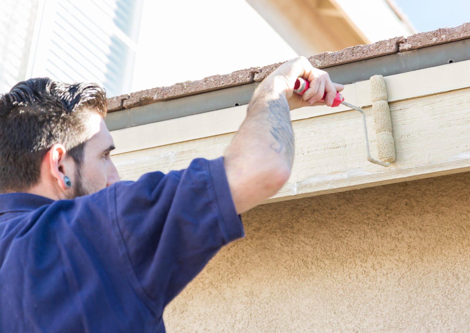 A painter uses a small roller to apply beige paint to exterior wood trim on a residential home.