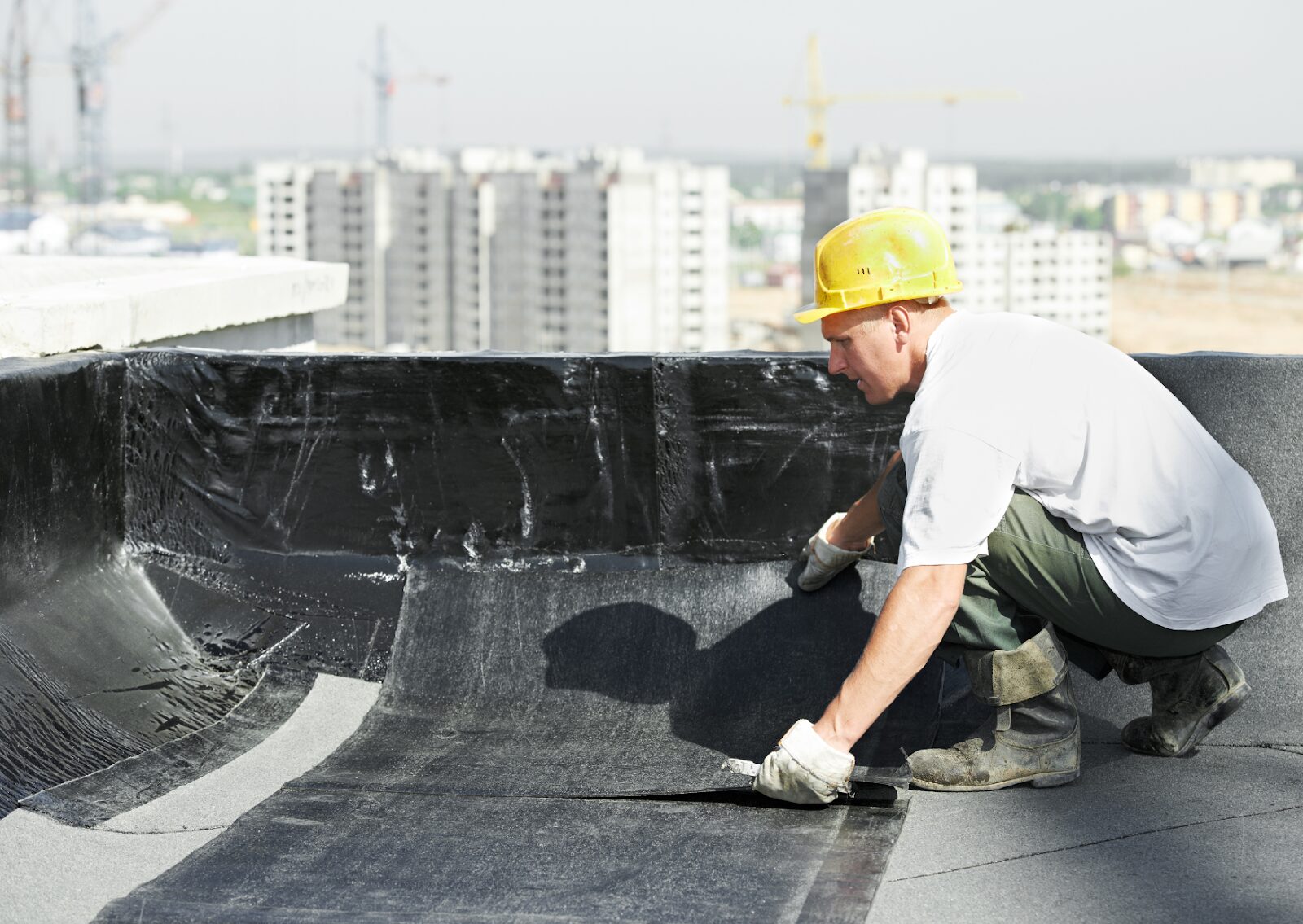 Construction worker installing roofing material on a flat commercial roof with city buildings in background.