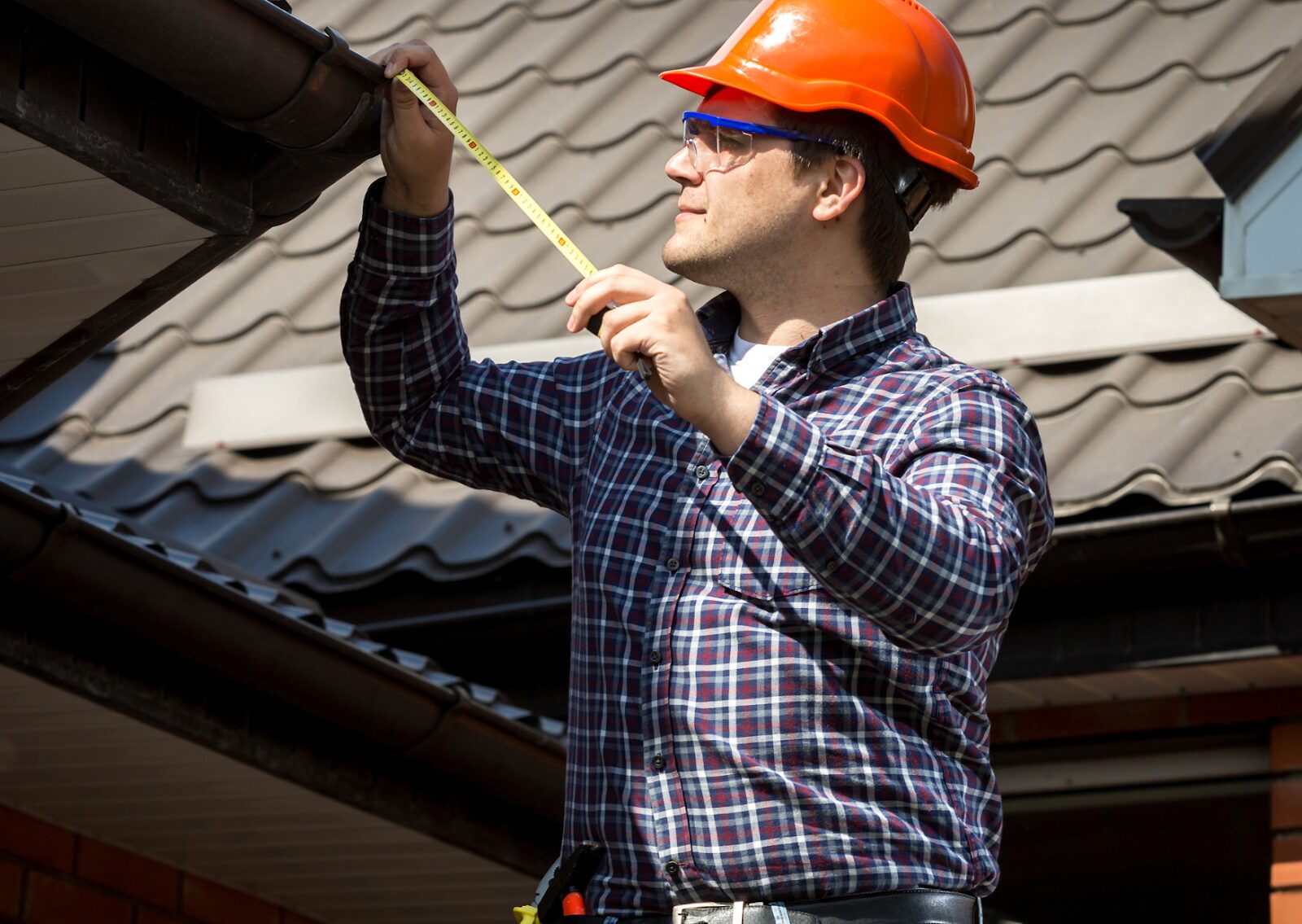 Construction worker in hard hat measuring a roof gutter with a tape measure during roof inspection.