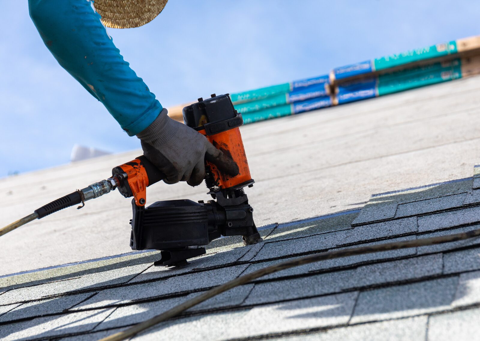Contractor using nail gun on asphalt shingles during roof repair for homes under clear blue sky.