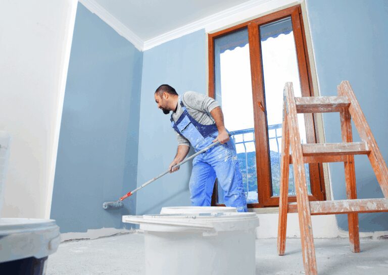 House painting professionals are applying blue paint to an interior wall with a roller next to a window.