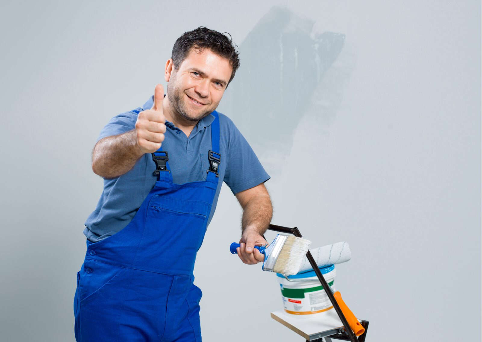 Smiling painter in blue overalls giving a thumbs up while holding a paintbrush in front of a painted wall.