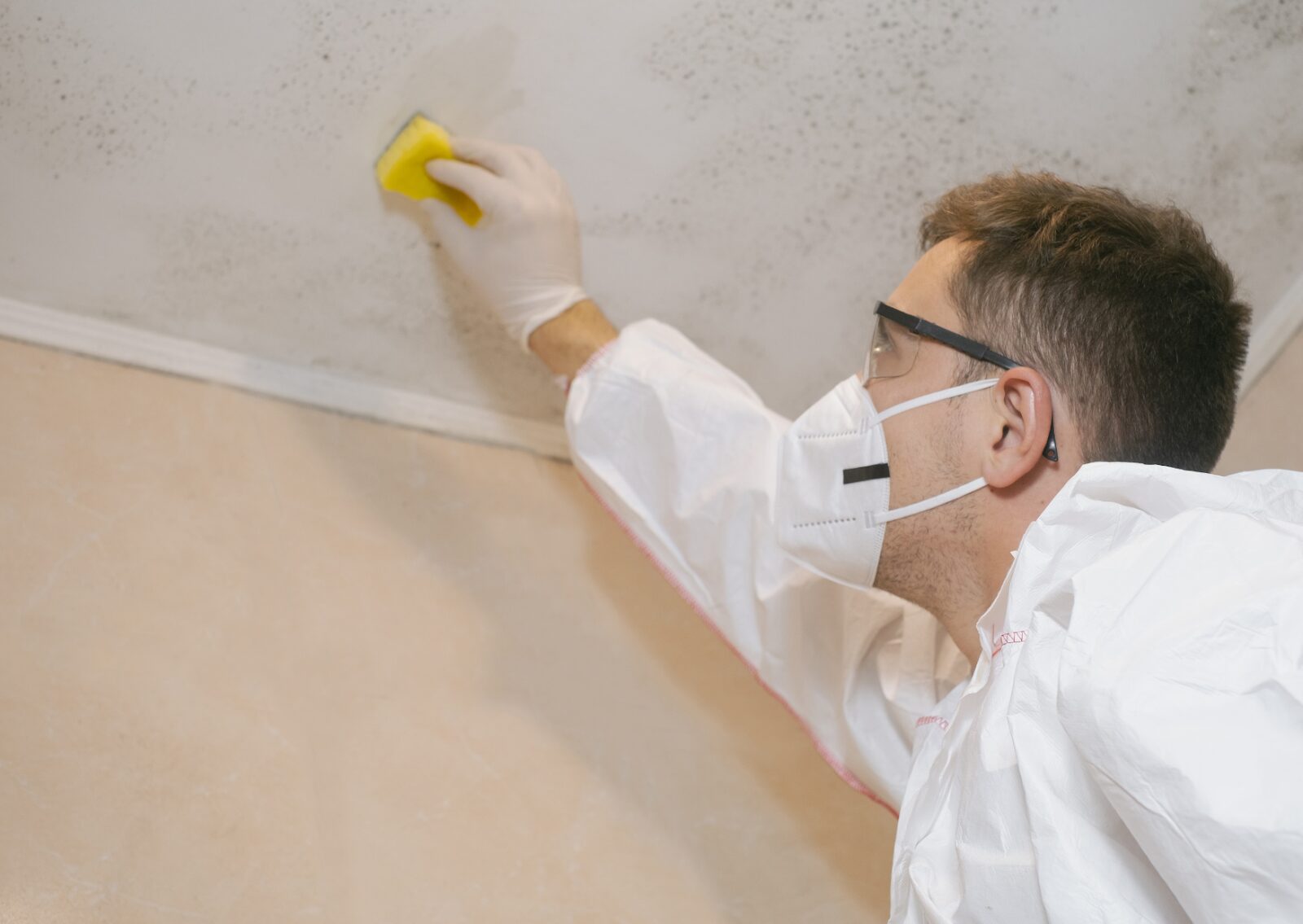 Worker in protective gear cleaning mold from ceiling as part of surface preparation before painting.