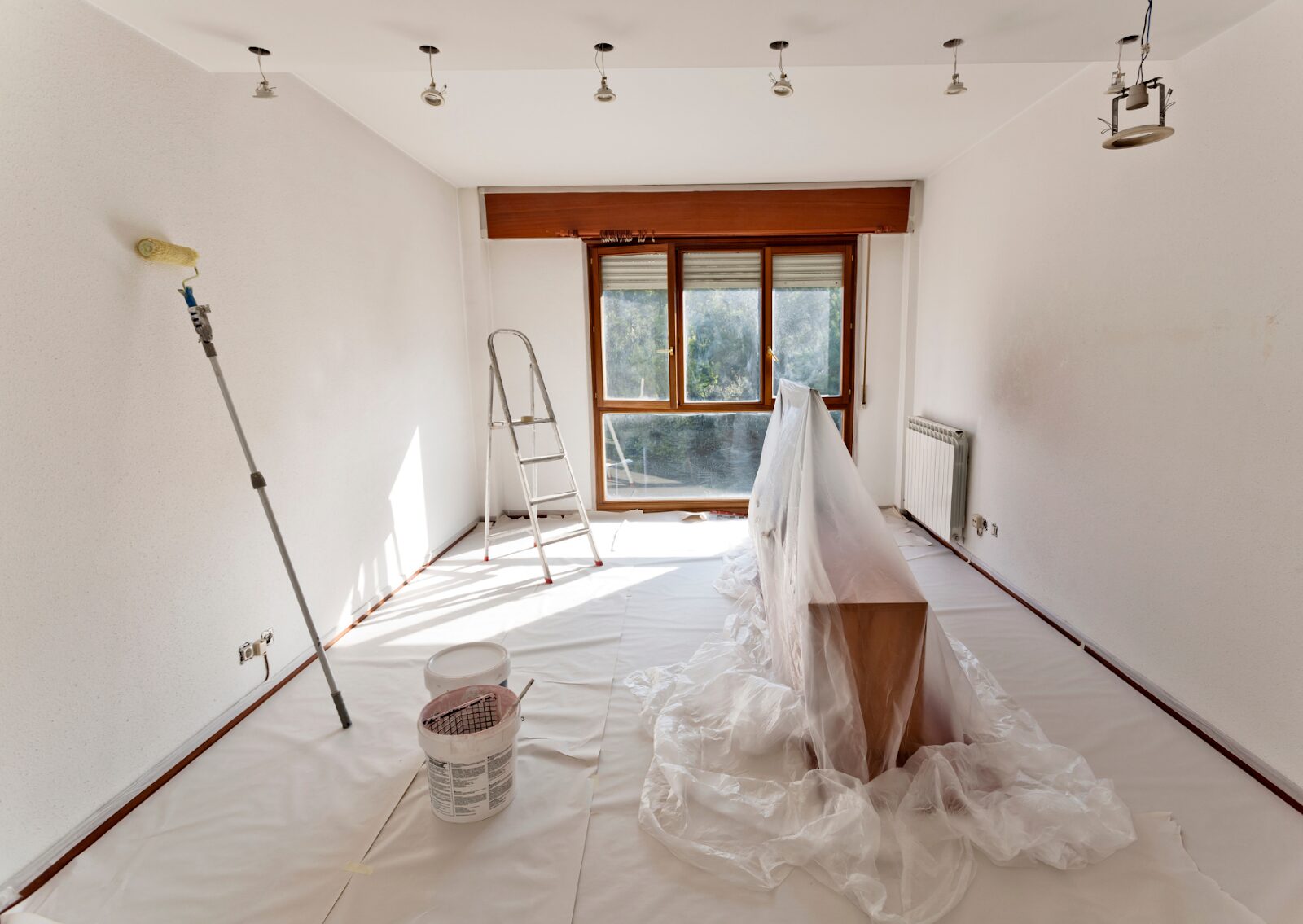 Room prepared with plastic sheets, ladder, and tools for surface preparation for painting interior walls.