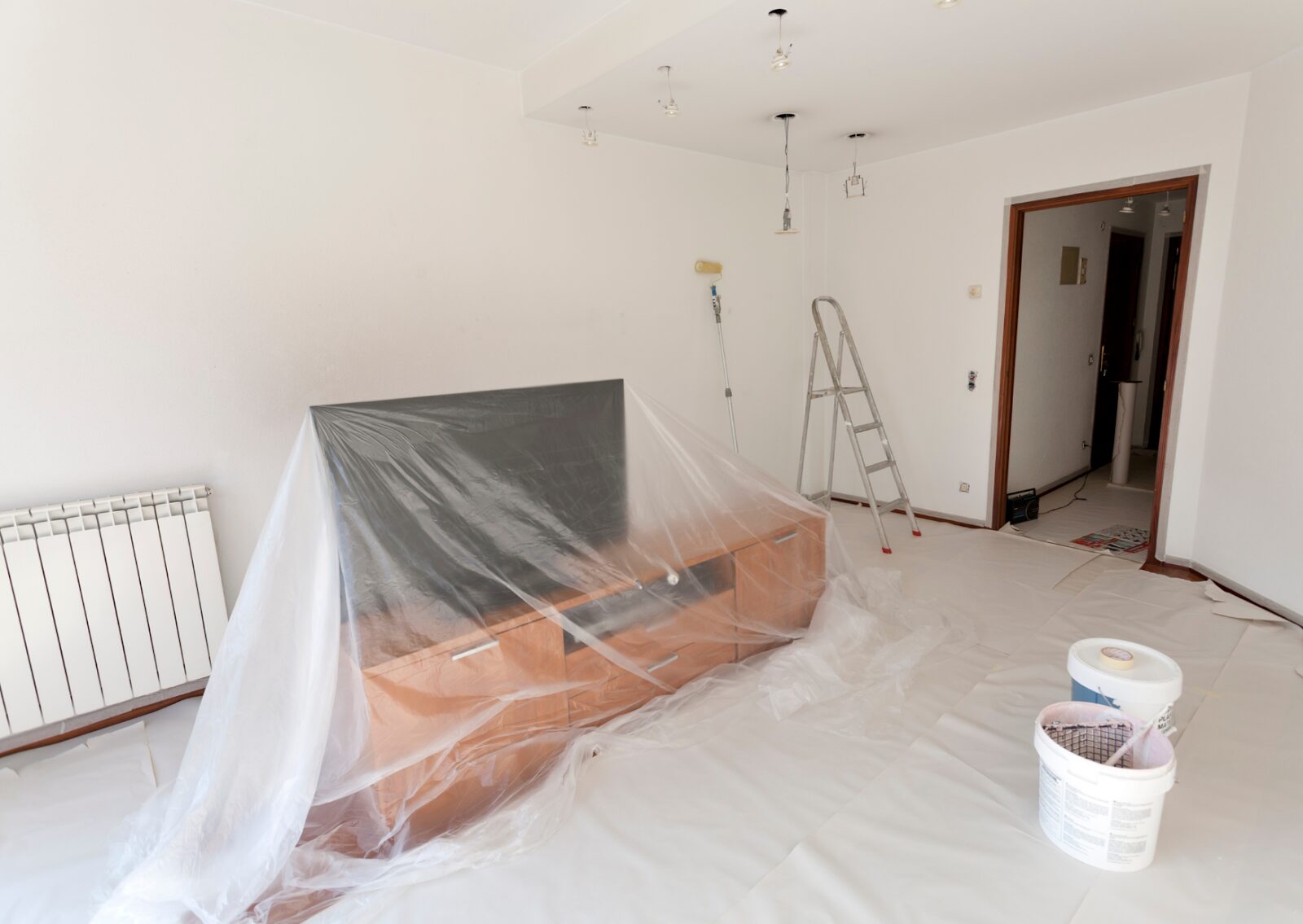 Living room prepared for painting with furniture covered in plastic, paint buckets, ladder, and roller on site.