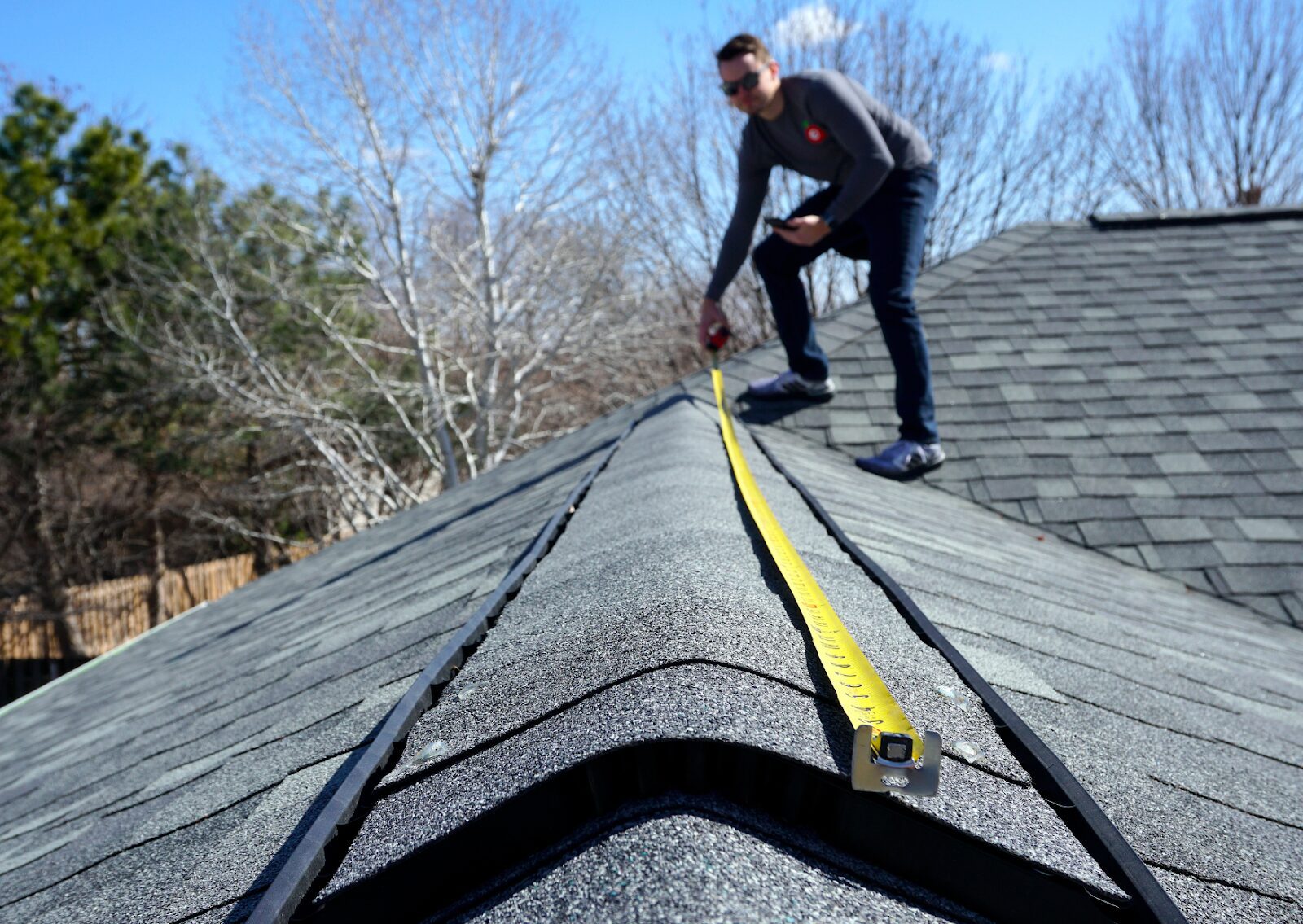 Contractor measuring rooftop ridge with tape measure during residential roofing inspection on a sunny day.